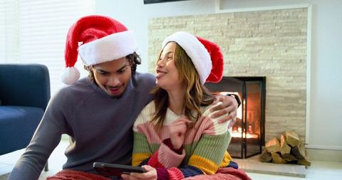 Couple wearing santa hats cozy by fireplace sharing tablet and laughing together for holiday warmth