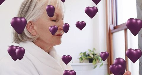 Senior woman relaxing in white robe by window with floating purple hearts for wellness