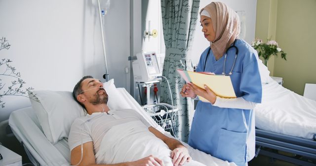 Female Muslim nurse wearing a hijab talking to a male patient lying in a hospital bed. She is holding a medical clipboard and has a professional appearance. Image can be used to depict caring in healthcare services, diverse medical workforce or nursing.