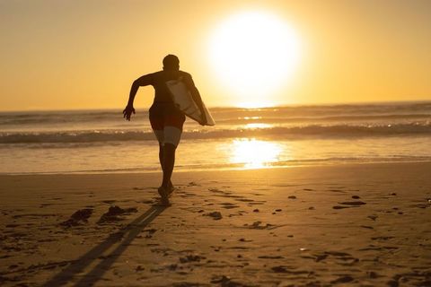 African American Surfer Running Toward Ocean at Golden Hour Carrying Shortboard Backlit