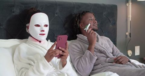 Couple enjoying at-home spa night wearing led face mask and using facial roller in bed