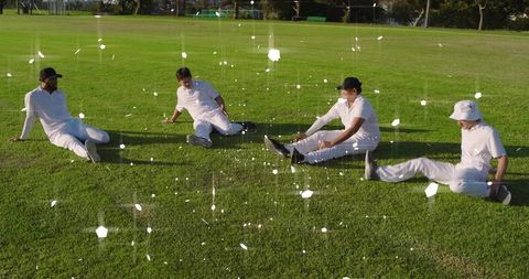 Cricket teammates resting on mown outfield wearing white kit and caps in sunny park