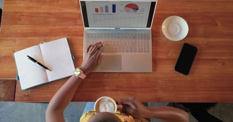 Woman working on laptop in cafe holding cappuccino with smartwatch, notebook and charts