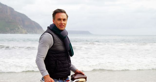 Man wearing scarf and vest standing on a beach with mountains in the background. The man looks relaxed while taking in the scenic view of the ocean. This image is suitable for themes related to leisure, travel destinations, outdoor activities, and enjoying nature.