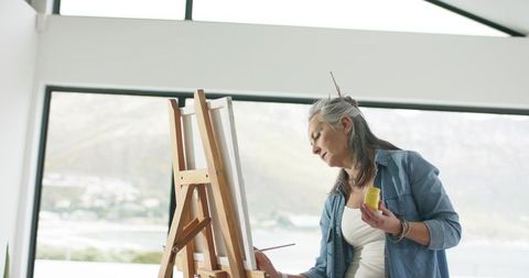 Mature woman painting on canvas in bright studio holding yellow paint jar