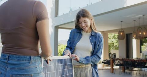 Two women installing portable solar panel on patio, smiling, sustainable home energy
