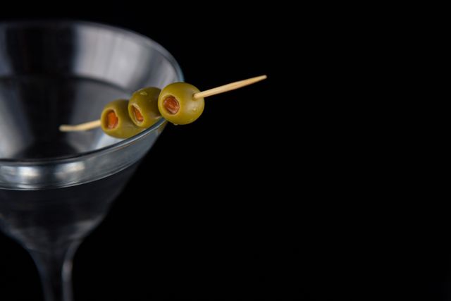Close-up of a martini glass with three green olives on a skewer against a black background. Perfect for use in bar and restaurant menus, cocktail recipes, party invitations, and beverage advertisements.