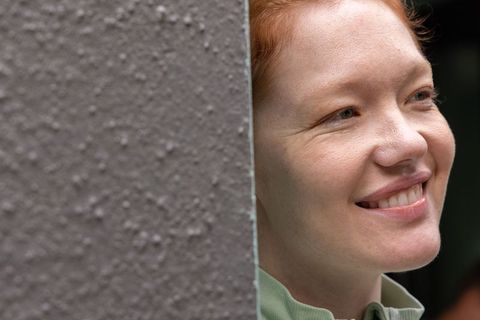 Smiling redhead woman peeking around textured wall, enjoying calm outdoor moment