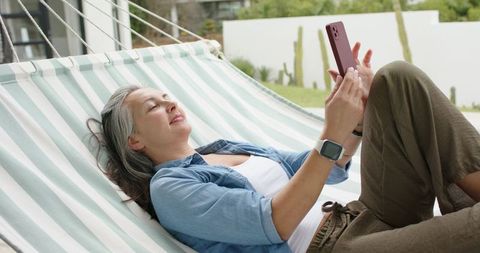 Mature woman smiling while relaxing on striped hammock in backyard using red smartphone