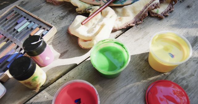 Brightly colored paint containers and pastels spread on a wooden table offering various art supplies. Suitable for concepts of creativity, arts and crafts, and artistic workspace. Ideal for use in educational content, art classes, DIY projects promotions, and creative workshops advertisements.