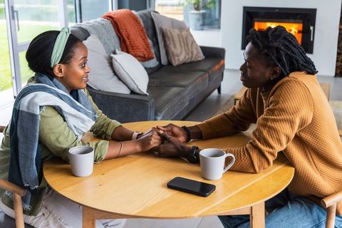 African american couple holding hands and talking at sunlit living room table by fireplace