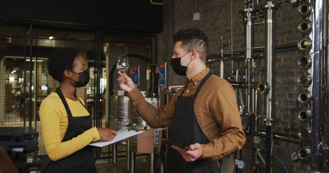 Team analyzing wine in a modern brewery or distillery. Both wearing face masks for safety, indicating a contemporary setting with an industrial vibe. Ideal for articles about workplace safety, brewing industry, and team collaboration.
