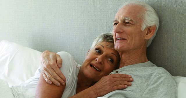 This image shows a senior couple embracing and relaxing in bed, conveying themes of love, togetherness, and a happy retirement. They appear content and comfortable, highlighting a moment of intimacy and affection. This image can be used for advertisements or articles on senior living, retirement planning, health and wellness, or relationships among elderly couples.