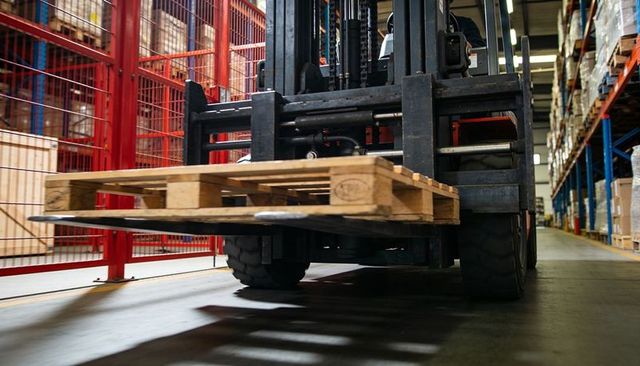 Forklift transporting wooden pallet through warehouse aisle with red safety barrier and shelving