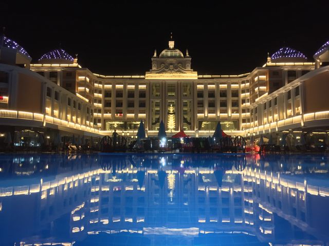 Luxurious grand hotel beautifully lit at night with its reflection in the calm pool water. The tall and elegant architecture, with multiple floors, windows lit, and intricate design details, creates a captivating view suitable for travel blogs, resort advertisements, and hospitality industry promotions emphasizing elegance and luxury.