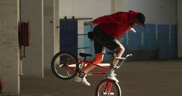 Bicycle rider performing thrilling stunts indoors in an empty warehouse. Red hoodie suggests vibrant activity. Perfect for use in urban sports advertisements, extreme sports magazines, or fitness promotion materials.