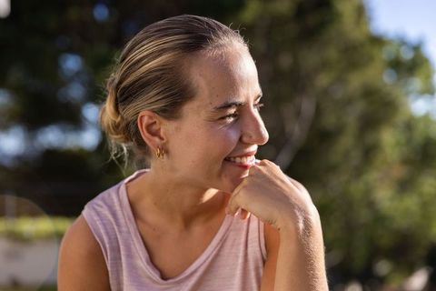 Smiling woman resting chin on hand in sunlit garden wearing light-pink top and gold hoops