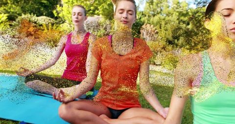 Women meditating on yoga mats outdoors in sunlit park surrounded by golden glitter energy