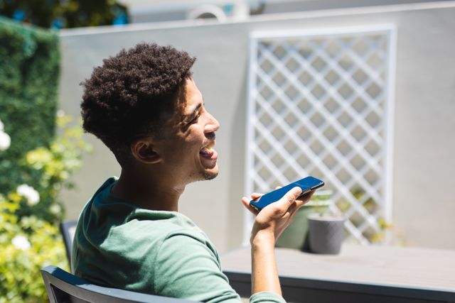 Side view of happy hispanic man talking on mobile phone through speaker at back yard on sunny day. unaltered, people, happiness, telecommunication and technology concept.