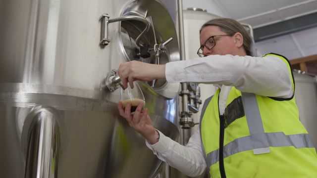 Employee sampling beer directly from fermentation tank wearing a high visibility vest. Ideal for illustrating articles on brewery processes, quality control in brewing, and craft beer production. Could be used for business and industrial brochures, showcasing behind-the-scenes of brewing operations or marketing materials for breweries.