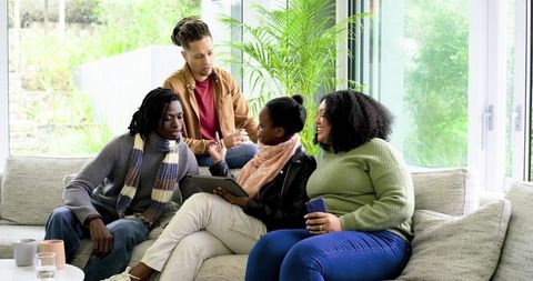 Multicultural friends sitting on sofa sharing tablet and laughing in bright living room