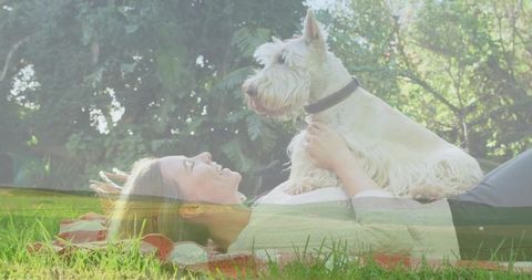 Smiling woman lying on picnic blanket holding white terrier under sunlit park trees
