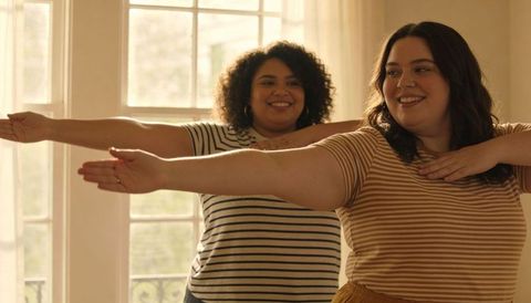 Smiling women practicing synchronized arm stretch in sunlit home studio wearing striped tops