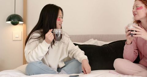 Diverse friends sharing coffee and chatting on bed in cozy pastel loungewear