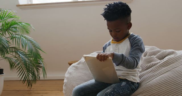 Young boy engaging with a digital tablet while sitting on a bean bag in a home environment. Ideal for use in content about children's technology use, online learning environments, modern family life, and home education. Highlights blend of comfort and technology in a domestic space.