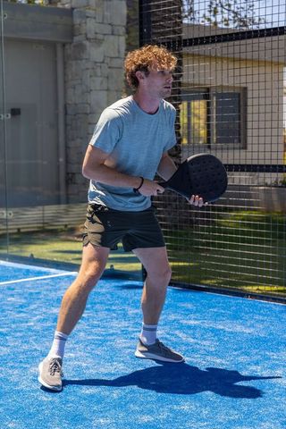 Curly-haired athlete preparing serve on blue outdoor padel court holding paddle ready