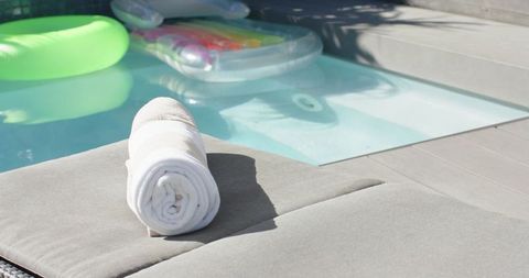 Rolled white towel resting on gray lounger by pool with inflatable floats and sunlight