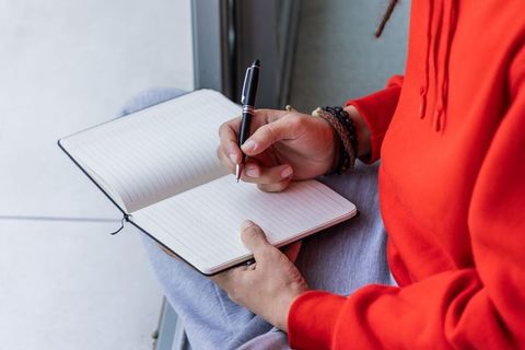 Man writing in lined notebook wearing red hoodie and beaded bracelets by sunlit window