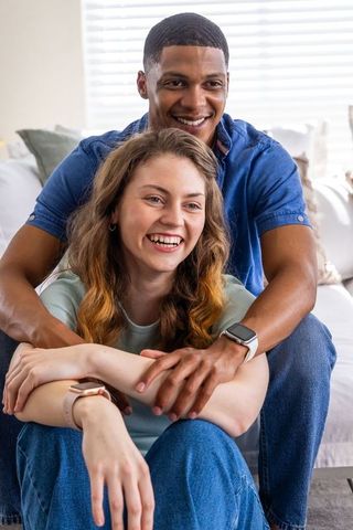Interracial couple laughing and embracing on couch showing smartwatches and cozy home life