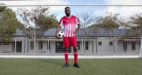 Adult soccer player standing in goal holding soccer ball in red uniform on artificial turf