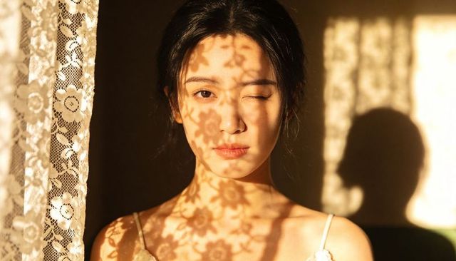 Asian woman winking behind lace curtain, floral shadows across face and camisole, sunlit