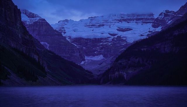Blue dusk glacier massif reflecting over calm alpine lake with conifer valley and snow at twilight