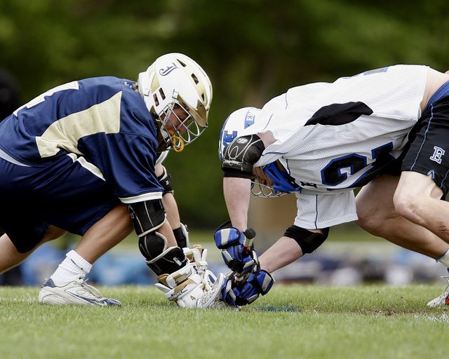 Lacrosse players crouching and competing for ball during a face-off on green field. Use for content related to sports, competition, teamwork, athletic events, youth sports, or lacrosse tutorials.