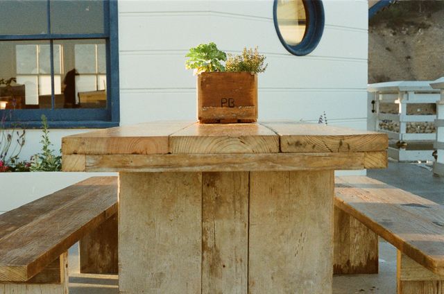Rustic wooden picnic table inviting relaxation in a sunlit outdoor setting. Small plant box on table enhances natural, laid-back atmosphere. Ideal for articles on outdoor décor, sustainable furniture, casual dining experiences, or nature-focused relaxation tips.
