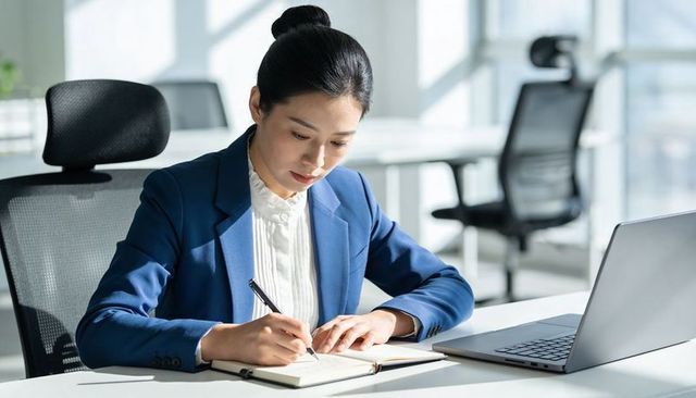 Asian businesswoman writing notes at bright modern office desk with laptop, planning