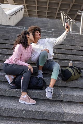 Diverse women friends in athletic wear taking selfie on concrete steps during outdoor fitness break