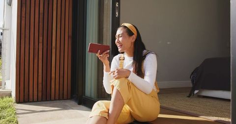 Sunlit asian woman sitting on doorstep laughing and holding smartphone in yellow overalls