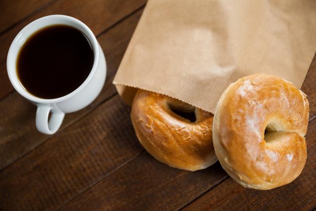 This photo shows a steaming cup of coffee alongside two fresh bagels partially inside a paper bag on a rustic wooden table. Ideal for use in breakfast-themed marketing materials, cafe menus, food blogs, and advertisements focusing on morning routines and simple pleasures.