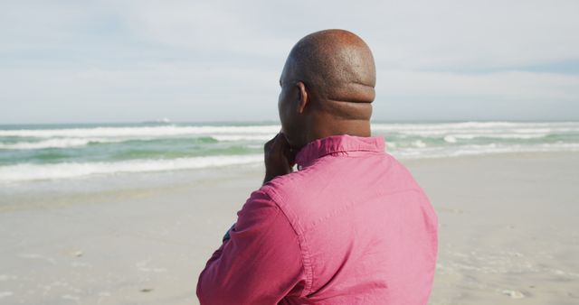 This image depicts a man standing on a beach, gazing thoughtfully at the ocean waves. Ideal for use in portrayals of solitude, contemplation, and relaxation. Suitable for mental health articles, travel promotions, and lifestyle blogs highlighting peaceful moments and personal reflections.
