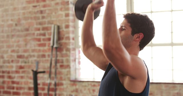 A young Caucasian man is lifting a dumbbell in a gym with brick walls, with copy space. His focus on fitness highlights the importance of regular exercise for maintaining health and well-being.