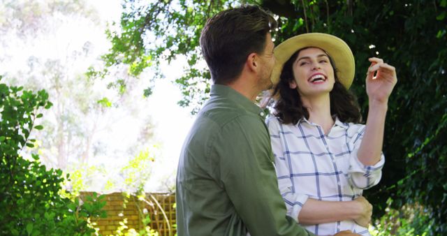Young couple spending time together in a lush garden, woman enjoying moment wear summer hat while man besides feeling happy and relaxed. Ideal for romance, leisure, outdoor activities, lifestyle blogs, and nature-related content.