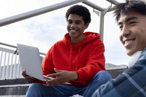 Teen friends studying together on campus steps sharing laptop smiling and wearing hoodies