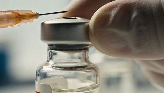 Preparing vaccine vial with gloved hand stabilizing stopper, syringe needle approaching