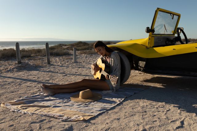 Caucasian man sitting on beach by beach buggy at sundown playing guitar. beach stop off on summer holiday road trip.
