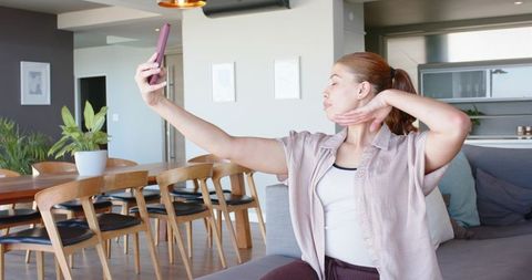 Young woman taking selfie on sofa with smartphone in modern open-plan apartment