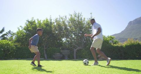 African american father and son playing soccer on sunny backyard lawn, family sport